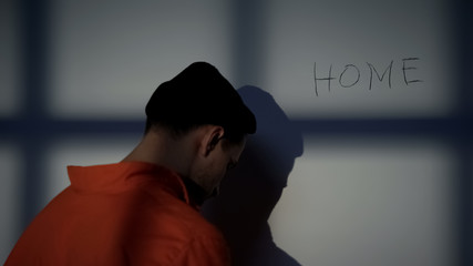Imprisoned male writing Home on cell wall, feeling homesick, hope for freedom