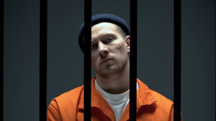 Dangerous criminal with scared face looking at camera behind prison bars