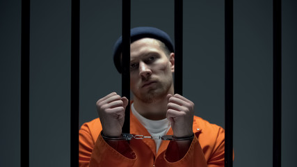 Dangerous arrogant prisoner standing behind bars and showing handcuffed hands