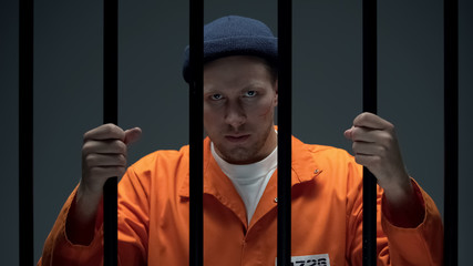 Angry Caucasian prisoner with scared face looking through cell bars, danger