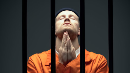 Imprisoned man in despair praying behind cell bars, feeling guilty about crime