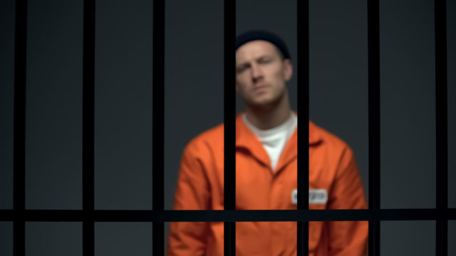 Blurred dangerous prisoner standing behind prison bars, justice, law and order