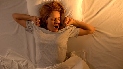 Attractive lady awakening in bed, yawning and stretching arms, happy morning