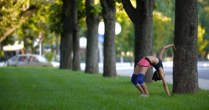 Beautiful Caucasian girl of seven years in sporty clothes practicing gymnastic exercises on the grass in a city park on a sunny summer day. Slow motion