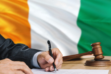 Judge writing on paper in courtroom with Cote D'Ivoire flag background. Wooden gavel of equality...