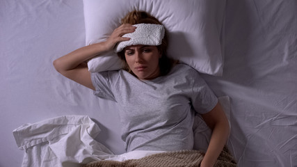 Upset woman with compress lying in bed, suffering from migraine and nausea
