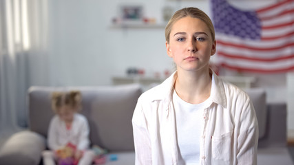 Fototapeta premium Sad single mother looking at camera daughter playing on sofa against flag of USA