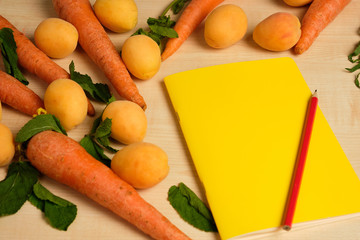 Yellow notepad with red pencil is surrounded by apricots and carrots on a wooden table