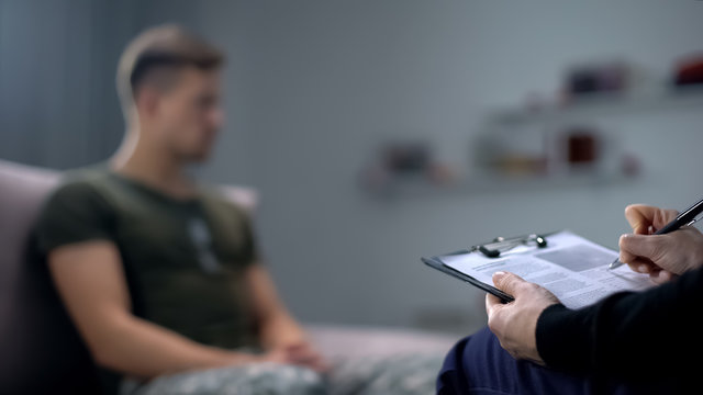 Psychologist making notes during therapy session with sad male soldier, PTSD
