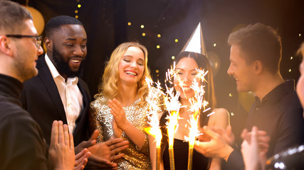 Friends congratulating pretty woman in party hat holding birthday cake sparklers