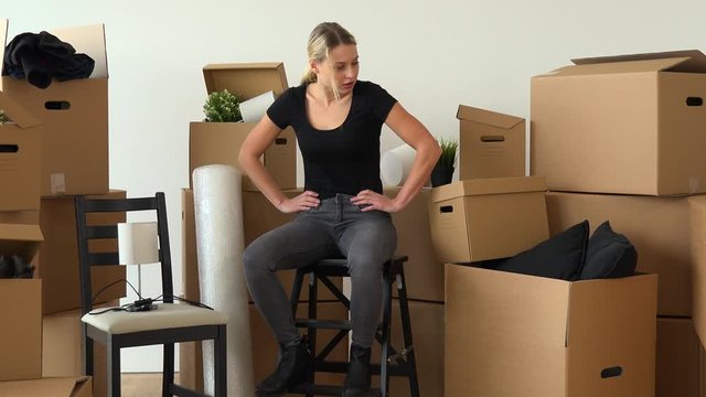 A moving woman sits on a chair in an empty apartment and acts irritated, surrounded by cardboard boxes