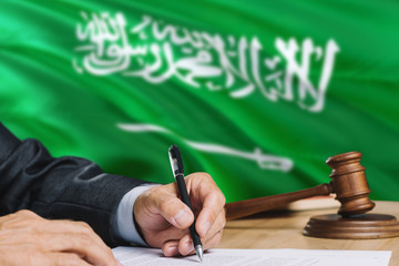 Judge writing on paper in courtroom with Saudi Arabia flag background. Wooden gavel of equality theme and legal concept.