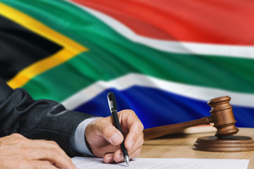Judge writing on paper in courtroom with South Africa flag background. Wooden gavel of equality...