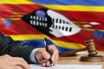 Judge writing on paper in courtroom with Swaziland flag background. Wooden gavel of equality theme and legal concept.