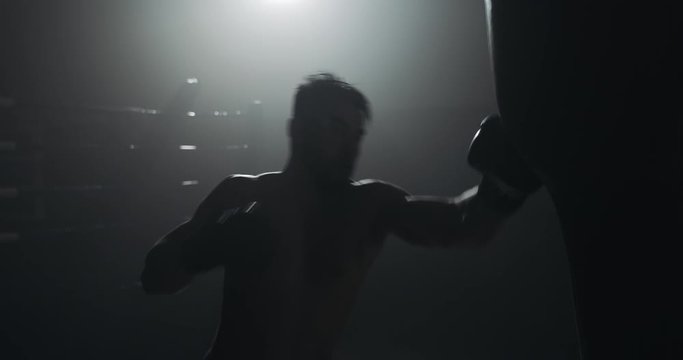 Silhouette boxer man hits punching bag in dark gym. Close up fighter workout. Male boxer exercising in dark. Kickboxer silhouette training.
