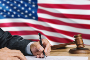 Judge writing on paper in courtroom with United States flag background. Wooden gavel of equality theme and legal concept.