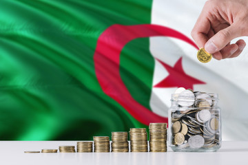 Business man holding coins putting in glass, Algeria flag waving in the background. Finance and...