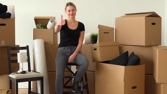 A happy moving woman sits on a chair in an empty apartment and shows a thumb up to the camera with a smile, surrounded by cardboard boxes
