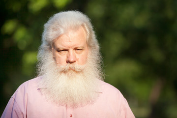 Abstracted bearded senior man in casual wear looks aside, standing against blur green natural background in city park at sunny day.