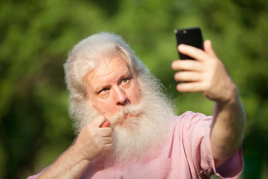 Bearded senior man in casual wear making selfie on smartphone and showing fig sign, standing against blur green natural background in city park at sunny day.