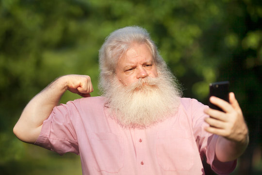 Funny bearded senior man with severe look making strong gesture and taking selfie on smartphone, standing against blur green natural background in city park at sunny day.