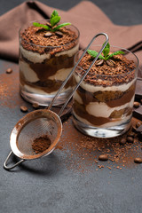 Classic tiramisu dessert in a glass cup, strainer and pieces of chocolate on dark concrete background