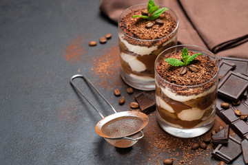 Classic tiramisu dessert in a glass cup, strainer and pieces of chocolate on dark concrete background