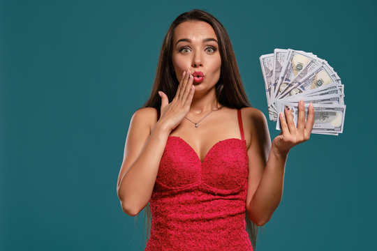 Brunette girl with a long hair, wearing a sexy red dress is posing holding a fan of hundred dollar bills against a blue background.