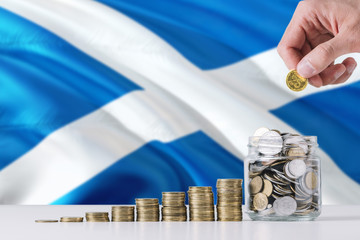 Business man holding coins putting in glass, Scotland flag waving in the background. Finance and business concept. Saving money.