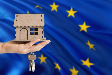 Woman hand holding wooden house and keys with hand. European Union flag with concept of rent,...