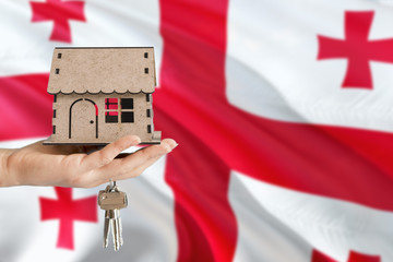 Woman hand holding wooden house and keys with hand. Georgia flag with concept of rent, purchase, insurance, building real estate, eco house.