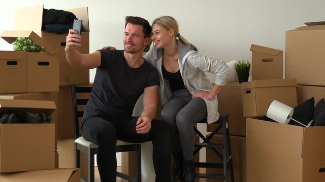 A moving couple sits on chairs and takes selfies with a smartphone in an empty apartment, surrounded by cardboard boxes