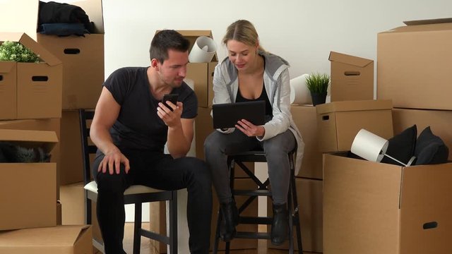 A smiling moving couple sits on chairs and works on a tablet and a smartphone in an empty apartment, surrounded by cardboard boxes