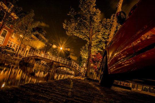 Delft / Netherlands - 05.21.2019: Night shot of a red car with a bridge over the canal in the background