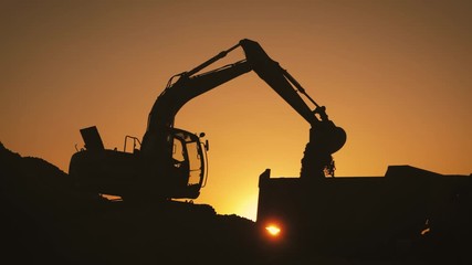 Silhouette of an excavator that loads sand into a truck at sunset. Concept construction and heavy industry, machine will be used in heavy industry business. Slow motion footage.