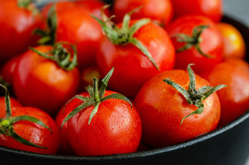 Red cherry tomatoes with water droplets in a black ceramic plate close-up on a dark concrete background.