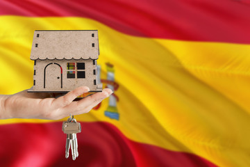 Woman hand holding wooden house and keys with hand. Spain flag with concept of rent, purchase, insurance, building real estate, eco house.