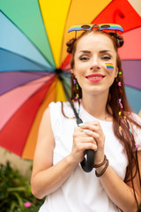 Happy lesbian with lgbt flag painted on her face posing with rainbow umbrella.
