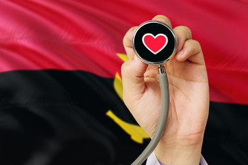 Doctor holding stethoscope with red love heart. National Angola flag background. Healthcare system concept, medical theme.
