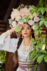 Brunette girl in a white ukrainian authentic national costume and a wreath of flowers is posing against a green yard. Close-up.