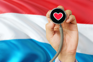 Doctor holding stethoscope with red love heart. National Luxembourg flag background. Healthcare system concept, medical theme.