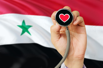 Doctor holding stethoscope with red love heart. National Syria flag background. Healthcare system concept, medical theme.