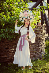 Obraz na płótnie Canvas Brunette girl in a white ukrainian authentic national costume and a wreath of flowers is posing against a wooden wicker fence.