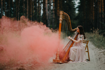 Woman harpist sits at forest and plays harp against a background of pines and smoke.