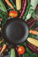 Creative layout made of corn, tomatoes, zucchini, carrot, beetroot, garlic, bean and black plate on dark background. Food concept. Flat lay, top view. Free space for your text.