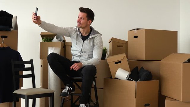 A moving man sits on a chair in an empty apartment and takes selfies, surrounded by cardboard boxes