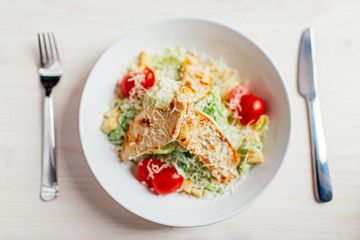 Fresh healthy caesar salad with chicken on white wooden table with fork and knife. Top view. Selective focus.