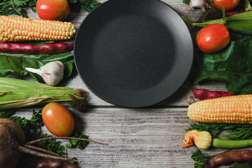 Assorted fresh vegetables on wooden background. Corn, tomatoes, zucchini, carrot, beetroot, garlic, bean and black plate. Food concept. Flat lay, top view, copy space