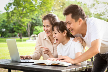 Happy asian family,father,mother,daughter enjoying,smiling using laptop computer in park,parents and child girl having fun playing game or watching  video,movie,social networks,media addiction concept