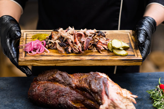 Grill restaurant kitchen. Chef hands in black cooking gloves holding smoked pulled pork with pickles on wooden board.